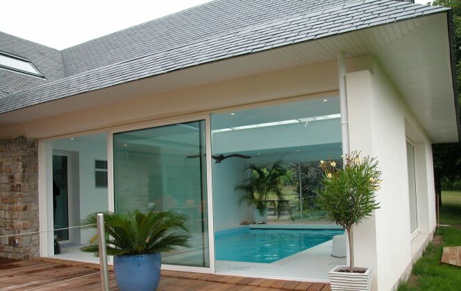 Piscine intérieure entourée de carrelage blanc, avec baie vitrée et terrasse en bois © L'Esprit Piscine